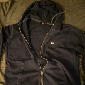 Mörkblå zip up från Lee - Säljer en mörkblå hoodie från Lee med dragkedja och huva. Den har en broderad logga på bröstet och snören vid huvan. Perfekt för en avslappnad stil. Hoddien är i mycket bra skick och storlek L