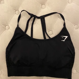 Svart sporttopp från Gymshark - Svart Gymshark sport-bh i storlek M. Med uttagbara pads. Välanvänd men har fortfarande mycket kvar att ge. Något noppig på banden och loggan är något sliten (se bild). Utöver det, inget annat att anmärka på. 94% nylon/6% elastan. 