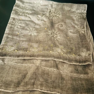 Beige halsduk/ sjal med blommigt mönster - Elegant beige halsduk/ sjal med ett subtilt blommigt mönster och små glittriga detaljer. Perfekt för att ge en touch av stil till din outfit. Lätt och luftig, idealisk för mildare väder.