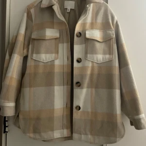 Beige och vit rutig kappa från H&M - Säljer en snygg beige och vit rutig overshirt från H&M. Jackan har stora knappar och två bröstfickor med lock. Perfekt för lager-på-lager-stil under hösten. 🧥