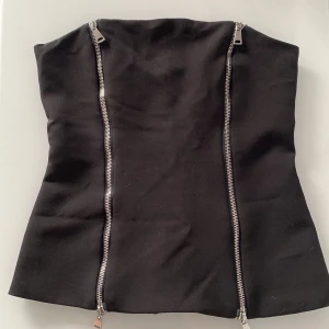 Svart korsett med dragkedjor - Snygg svart korsett med två dragkedjor framtill. Perfekt för en stilren och edgy look. Korsetten är axelbandslös och har en figurnära passform.