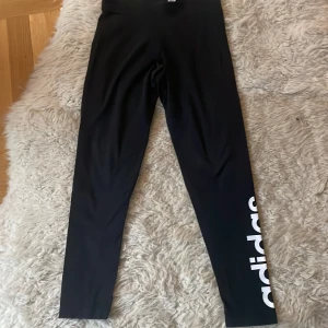 Svarta leggings från Adidas - Snygga svarta leggings från Adidas med vit logga längs benet. Perfekta för träning eller en avslappnad dag. Tillverkade i ett stretchigt material för bästa komfort.