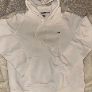 Vit hoodie från Tommy Jeans - Säljer en stilren vit hoodie från Tommy Jeans med klassisk logga på bröstet. Hoodien har en justerbar dragsko i huvan och en praktisk magficka. Använd en gång