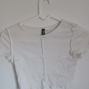 Vit croppad topp från Divided H&M - Säljer en vit croppad topp från Divided H&M. Toppen har korta ärmar och synliga sömmar som ger en trendig look. Perfekt för sommardagar eller som en del av en lager-på-lager outfit.