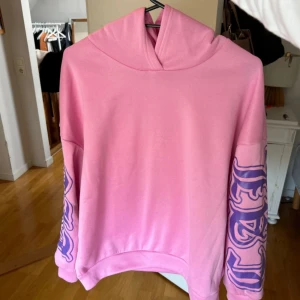 Rosa hoodie med tryck - Säljer en rosa hoodie med tryck i lila på ärmarna och ryggen. Hoodien har en stor och mysig huva och är perfekt för en avslappnad stil. Texten på ryggen ger en cool och unik touch. 