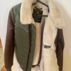 Grön och brun jacka från Alpha Industries - Säljer en FET grön och brun jacka från Alpha Industries med en mysig pälsfodrad insida och krage. Jackan har långa ärmar och en dragkedja framtill. Den har också en praktisk ficka med dragkedja på ärmen. Passar S/M