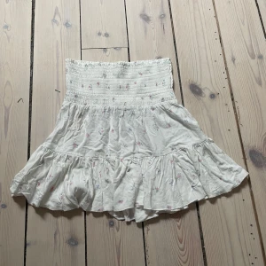 Kjol, blommig, vit från Lager 157 - Söt vit kjol med blommigt mönster i rosa och blått. Kjolen har en smockad midja. Den har en liten fläck som knappt syns (se bild 3)