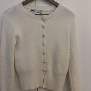 Vit kofta från Zara - Säljer en vit kofta från Zara med dekorativa knappar. Koftan har en rund halsringning och långa ärmar, perfekt för en basic men även fin och uppåiffande outfit. Passar bra till både jeans och kjol. Den passar även för xs och xxs. Tröjan är i ett super skick då den andvänds fåtal gånger och bär inga defekter .