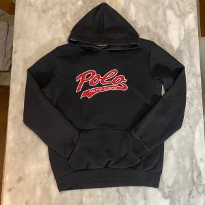 Svart hoodie från Ralph Lauren - Snygg svart hoodie från Ralph Lauren med röd och vit Polo-logga på bröstet. Perfekt för en avslappnad stil med en stor ficka framtill och en bekväm huva.