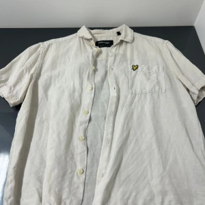 Beige kortärmad skjorta från Lyle & Scott - Säljer en stilren beige kortärmad skjorta från Lyle & Scott. Skjortan har en klassisk krage och en bröstficka med en broderad logotyp. Perfekt för en avslappnad stil. Skick 6/10 