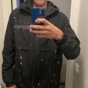 Svart vindjacka från Calvin Klein - Snygg svart vindjacka från Calvin Klein med dragkedja och huva. Perfekt för blåsiga dagar. Jackan har en stilren design med diskret logga på bröstet.