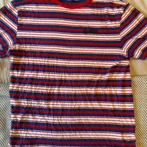 Randig t-shirt från Divided - Säljer en randig t-shirt från Divided i rött, blått och vitt. Den har korta ärmar och en rund halsringning med röd kant. 