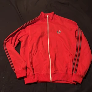 Röd track jacket från Fred Perry (STL L) - Snygg röd track jacket från Fred Perry med vita dragkedjor och klassisk logga på bröstet. Jackan har svarta ränder längs ärmarna och ribbade muddar vid ärmslut och nederkant. Perfekt för en sportig look!