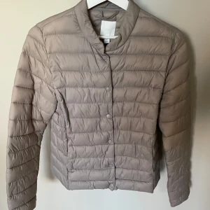 H&M gråbeige jacka, storlek 38 - *Snygg och lätt vadderad jacka i gråbeige nyans. *Jackan har en rak, figursydd modell med knappar fram.   *Den har en klassisk krage och långa ärmar.   *Perfekt för vår och höst. *Mycket sparsamt använd(2-3 gånger).  *Som ny. Storleken är mer som en S *Tvättad *Kommer från djurfritt och rökfritt hem