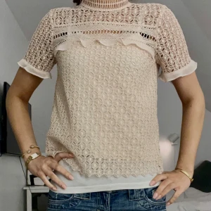 Beige spetsblus - Säljer en elegant beige spetsblus med korta ärmar och vackra spetsdetaljer. Blusen har en hög hals och volangdetaljer framtill. Perfekt för en stilren look.