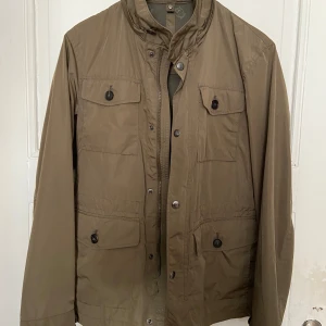Olivgrön vårjacka från Massimo Dutti - Snygg khaki färgad jacka från Massimo Dutti med dragkedja och knappar. Jackan har flera praktiska fickor och en avtagbar huva. Perfekt för höst och vår med sin stilrena design och funktionella detaljer. Jackan är som ny🤗