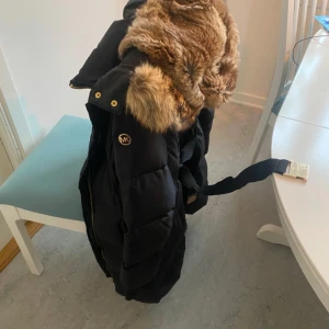 Marin blå dunjacka med äkta räv päls  från Michael Kors - Snygg lite tunnare dunjacka  från Michael Kors med päls på huvan. Kappan har en dragkedja och knappar i guld, samt ett bälte i midjan för en figursydd look. Perfekt för kalla dagar med sin varma och stilrena design. Ny pris 11 000kr Dragkedjan går inte att knäppa därav priset. Går att laga ev byta.
