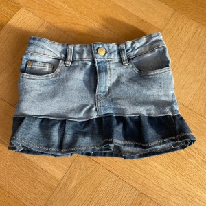 Jeanskjol med volang - Söt jeanskjol i ljusblå denim med volangdetalj längst ner. Kjolen har klassiska femficksdetaljer och en knapp framtill. Perfekt för en avslappnad stil.