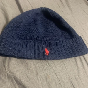 Mörkblå stickad mössa från Polo Ralph Lauren - Snygg mörkblå stickad mössa från Polo Ralph Lauren med röd broderad logotyp framtill. Perfekt för vinterns kyliga dagar, köptes från johnells för 999kr, buda gärna! 