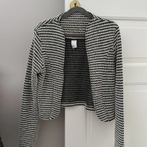 Svartvit mönstrad jacka från Vila - Snygg svartvit mönstrad jacka från Vila. Jackan har en öppen design utan knappar. Den har långa ärmar och en croppad passform som ger en modern look🤍
