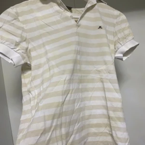 Randig pikétröja från J.Lindeberg - Säljer en stilren randig pikétröja från J.Lindeberg i beige och vit. Tröjan har korta ärmar och en klassisk krage. Perfekt för en avslappnad men ändå snygg look.