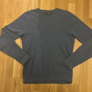 Blå stickad tröja från Dressmann - Säljer en stilren blå stickad tröja från Dressmann. Tröjan har en klassisk rund hals och långa ärmar, perfekt för kyligare dagar. Den är mjuk och bekväm, idealisk för både vardag och avslappnade tillfällen.