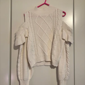 Vit stickad offshoulder tröja - Säljer en snygg vit stickad tröja med offshoulder-design och flätmönster. Tröjan har långa ärmar och ribbade kanter. Perfekt för en stilren look.