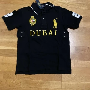 Cheif keef Ralph Lauren polo - Snygg svart pikétröja från Ralph Lauren med broderad Dubai-text och emblem på bröstet. Tröjan har korta ärmar och en klassisk krage med vita detaljer. Nummer 2 är broderat på ena ärmen. Perfekt för en stilren look. 9/10 skick oanvänd 
