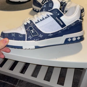 Blå och vita sneakers från Louis Vuitton - Snygga sneakers från Louis Vuitton i blått och vitt med monogrammönster. Skorna har en robust sula och klassisk snörning. Perfekta för en stilren look med en touch av lyx.