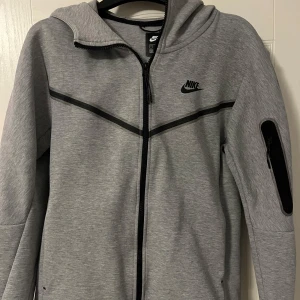 Nike tech - Säljer en grå hoodie från Nike med dragkedja och huva. Den har svarta detaljer och en ficka med dragkedja på ärmen. Perfekt för en sportig look eller avslappnade dagar.