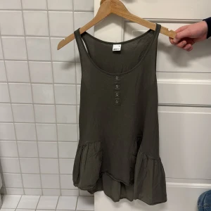 Olivgrön linnetopp från Gina Tricot - Säljer en olivgrön linnetopp från Gina Tricot i storlek XS. Toppen har knappar framtill och en volangdetalj vid nederkanten. Perfekt för en avslappnad stil.