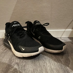 Svarta Nike Air Max 270 - Säljer ett par svarta Nike Air Max 270 sneakers med vit sula och synlig Air-enhet i hälen. Skorna har snörning och en stilren design med Nike-logga på sidan.