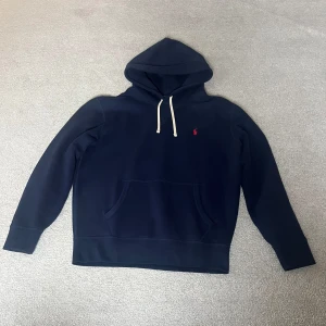 Mörkblå hoodie från Ralph Lauren - Säljer en stilren mörkblå hoodie från Ralph Lauren med en liten röd logga på bröstet. Den har en klassisk känguruficka och justerbar huva med vita snören. Perfekt för en avslappnad look. Använd bara några få gånger.