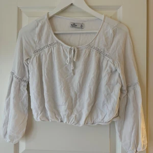 Vit blus från Hollister - Säljer en söt vit blus från Hollister med knytband framtill och spetsdetaljer på ärmarna. Blusen har långa ärmar, en lös passform och kroppad💗