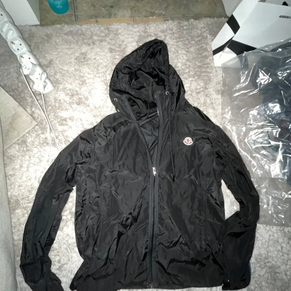 Snygg svart vindjacka från Moncler med huva och dragkedja framtill. Perfekt för blåsiga dagar och har en stilren design med Moncler-logga på bröstet. NFC funkar. Takit.
