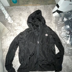 Svart Moncler Windbreaker - Snygg svart vindjacka från Moncler med huva och dragkedja framtill. Perfekt för blåsiga dagar och har en stilren design med Moncler-logga på bröstet. NFC funkar