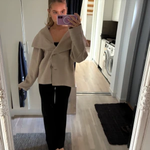 Beige kappa med bred krage - Säljer en stilren beige kappa med bred krage och knappar framtill. Jackan har en lös passform och långa ärmar, perfekt för höst och vår. Den är tillverkad i ett mjukt material som ger en elegant look.