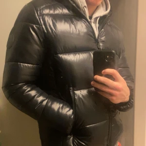 Svart pufferjacka från Save the Duck - Nytt skick 10/10, 100% Äkta vara. Snygg svart pufferjacka från Save the Duck med huva och dragkedja. Jackan är tillverkad i syntetmaterial och har en glansig finish. Perfekt för kyliga dagar med sin värmeisolerande Plumtech- material. 