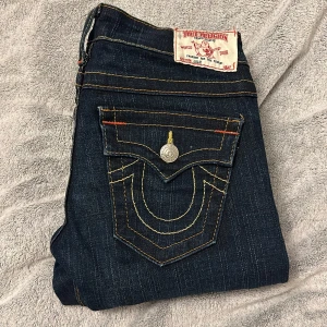 Mörkblå jeans från True Religion - Snygga mörkblå jeans från True Religion med ikoniska bakfickor och kontrastsömmar. De har en klassisk femficksdesign och knappar med märkets logga. Perfekta för en stilren look.