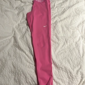 Rosa träningstights från Nike - Snygga rosa träningstights från Nike Pro med en elastisk midja och vit logga på benet. Perfekta för gymmet eller löprundan. De är designade för att sitta bekvämt och ge stöd under träning.