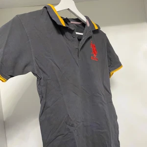 Grå pikétröja med gula detaljer - Säljer en stilren grå pikétröja med gula detaljer på krage och ärmslut. Tröjan har en broderad röd logotyp på bröstet och knappar vid halsen. Perfekt för en avslappnad stil.