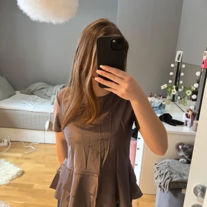 Grå topp från Atelier Capri - Säljer en elegant lila topp från Atelier Capri. Toppen har korta ärmar och en volangdetalj vid nederkanten som ger en fin siluett. Perfekt för en stilren look. Storlek M men passar mig som vanligt viss har xs-s