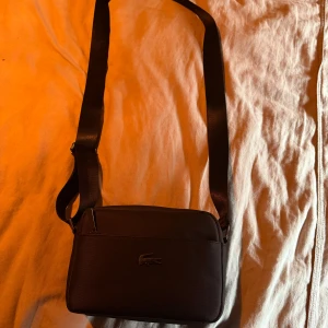 Svart axelväska från Lacoste - Snygg svart axelväska från Lacoste med justerbar rem. Väskan har en stilren design med Lacoste-loggan på framsidan. Perfekt för att bära dina nödvändigheter med stil.