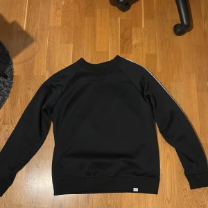 Svart tröja med vita detaljer från Les deux - Snygg svart tröja från Les deux med vita ränder längs ärmarna och logotyp. Tröjan har en klassisk passform med långa ärmar och rund halsringning. Perfekt för en stilren look. 