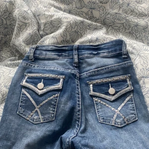 Blå jeans med broderade bakfickor - Snygga blå jeans med unika broderade detaljer på bakfickorna och en dragkedja framtill. De har en klassisk femficksdesign och en knappstängning. Perfekta för en casual look.