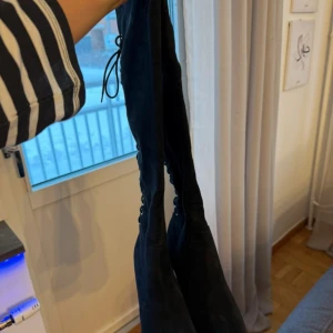 Svarta overknee stövlar med snörning  - Snygga svarta höga stövlar i mocka med snörning längs baksidan. Perfekta för att ge en stilren look med en touch av elegans. De har en klack som ger extra höjd och stil. Helt nya 