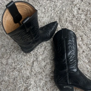 Svarta cowboy boots från Sendra - Snygga svarta cowboy boots från Sendra i skinn med dekorativa broderier. De har en spetsig tå och klassisk cowboy-stil. Perfekta för att ge din outfit en cool touch.