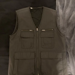 Mörkgrå quiltad väst med dragkedja - Säljer en mörkgrå quiltad väst med dragkedja . Västen har flera praktiska fickor med dragkedjor och lock. Västen passar streetwear. Storleken S men skulle säga att det bara är med tjocktröja då västen är rätt bred, annars utan  tjocktröja är storleken M. 
