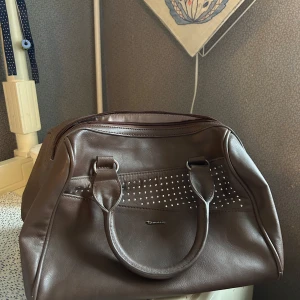 Brun handväska med nitar - Snygg brun handväska från Tamaris i syntetmaterial. Väskan har dekorativa nitar på framsidan och en praktisk dragkedja upptill. Perfekt för att bära dina dagliga nödvändigheter med stil.