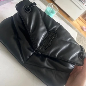 Svart quiltad axelväska från Steve Madden - Snygg svart axelväska från Steve Madden i quiltat syntetmaterial. Väskan har en kedjerem och en stilren logga framtill. Perfekt för att ge din outfit en trendig touch.